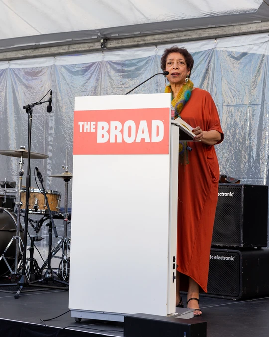 Woman with short curly hair, wearing a red dress, speaking at a podium with a sign for The Broad on front