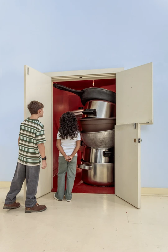 Two young children peeking into a red closet filled with oversized pots and pans